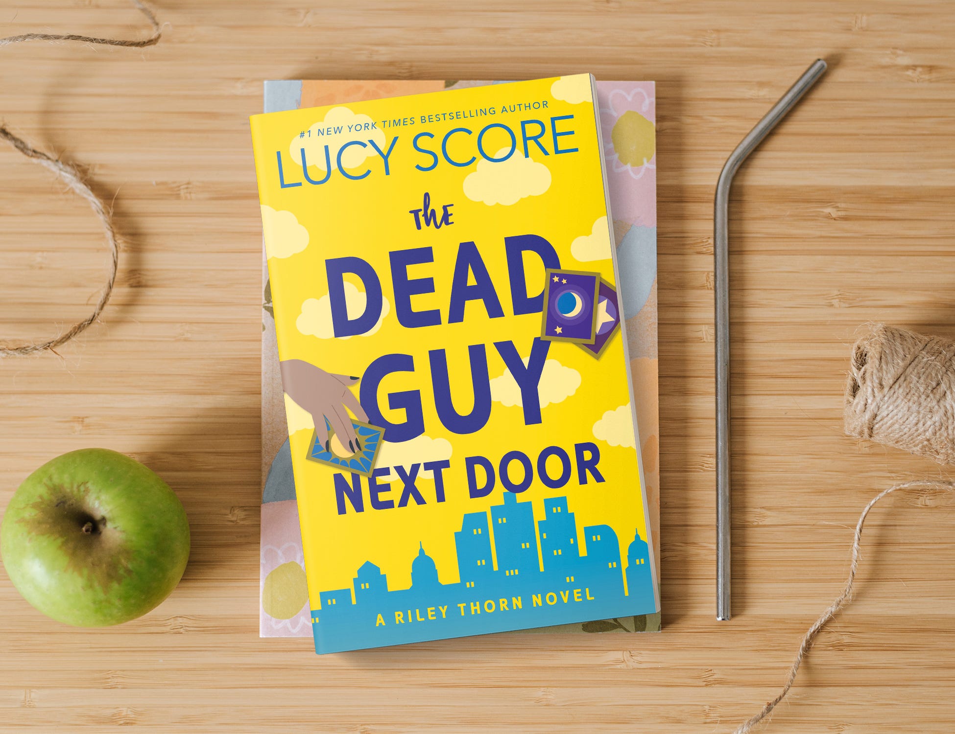 Image of The Dead Guy Next Door by Lucy Score book on a wood table next to a straw, a green apple, and string